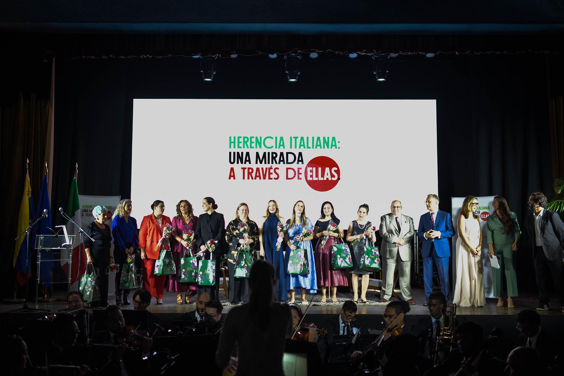 eredita italiana gruppo di persone su un palco con dietro un tabellone bianco e delle scritte verdi, nere e rosse, edavanti un'orhestra non illuminata 